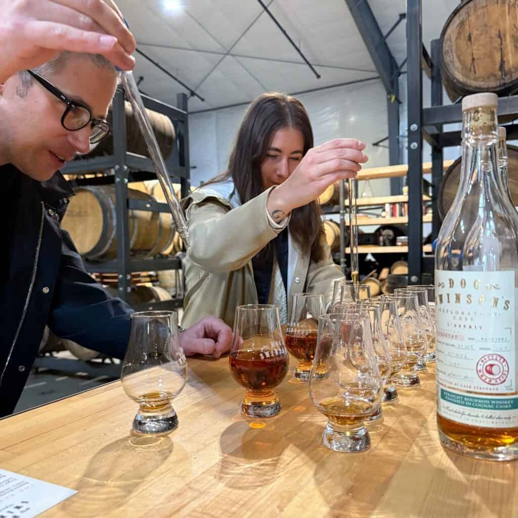 Dva lidé používají pipety k přidávání vody do sklenic whisky na dřevěném stole v palírně se sudy a lahví "Doktora Swinsona" v pozadí.