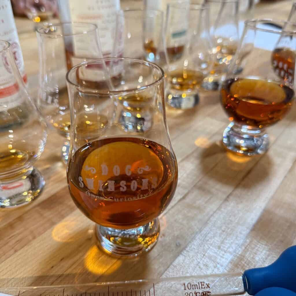 Několik sklenic jantarové tekutiny, pravděpodobně whisky, je uspořádáno na dřevěném stole s pipetou a odměrným válcem v popředí.