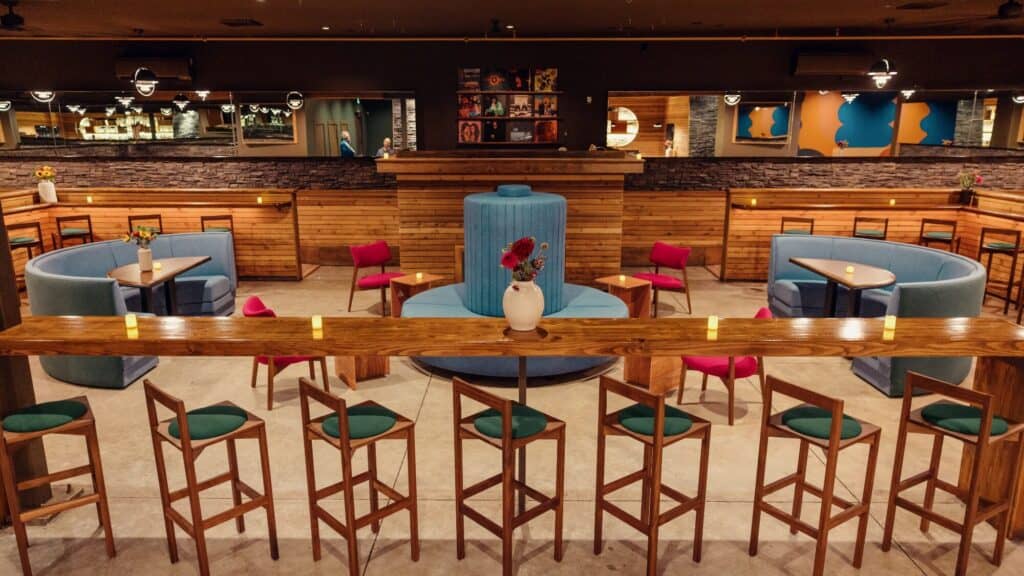 A modern bar with wooden high tables and chairs, round booths, pink chairs, a central blue fountain, and ambient lighting.
