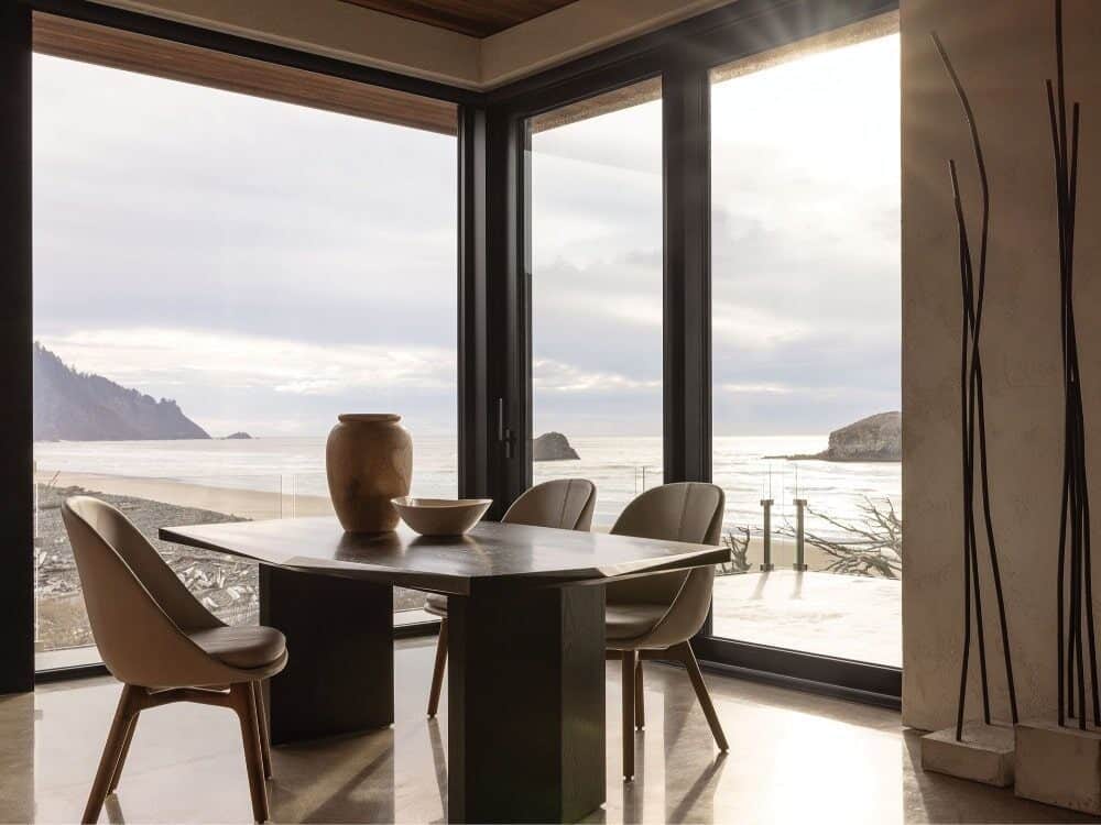 A modern dining area with a wooden table, chairs, and ceramic vase overlooks an Oregon beach through large floor-to-ceiling windows.