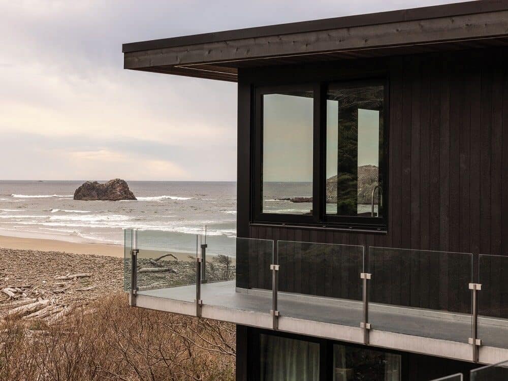 A modern house with dark wood siding and glass balcony overlooks an Oregon beach at Arch Cape, with a sandy shore, ocean views, and a large rock in the water under a cloudy sky.