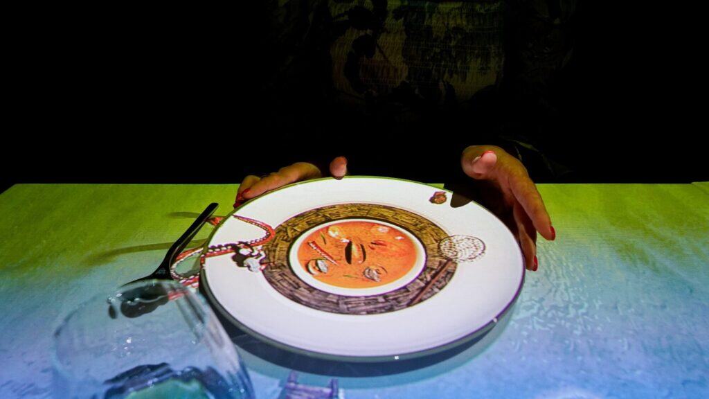 A person holds a plate with an illustrated soup design on it, next to a drinking glass and utensils on a light-colored table.