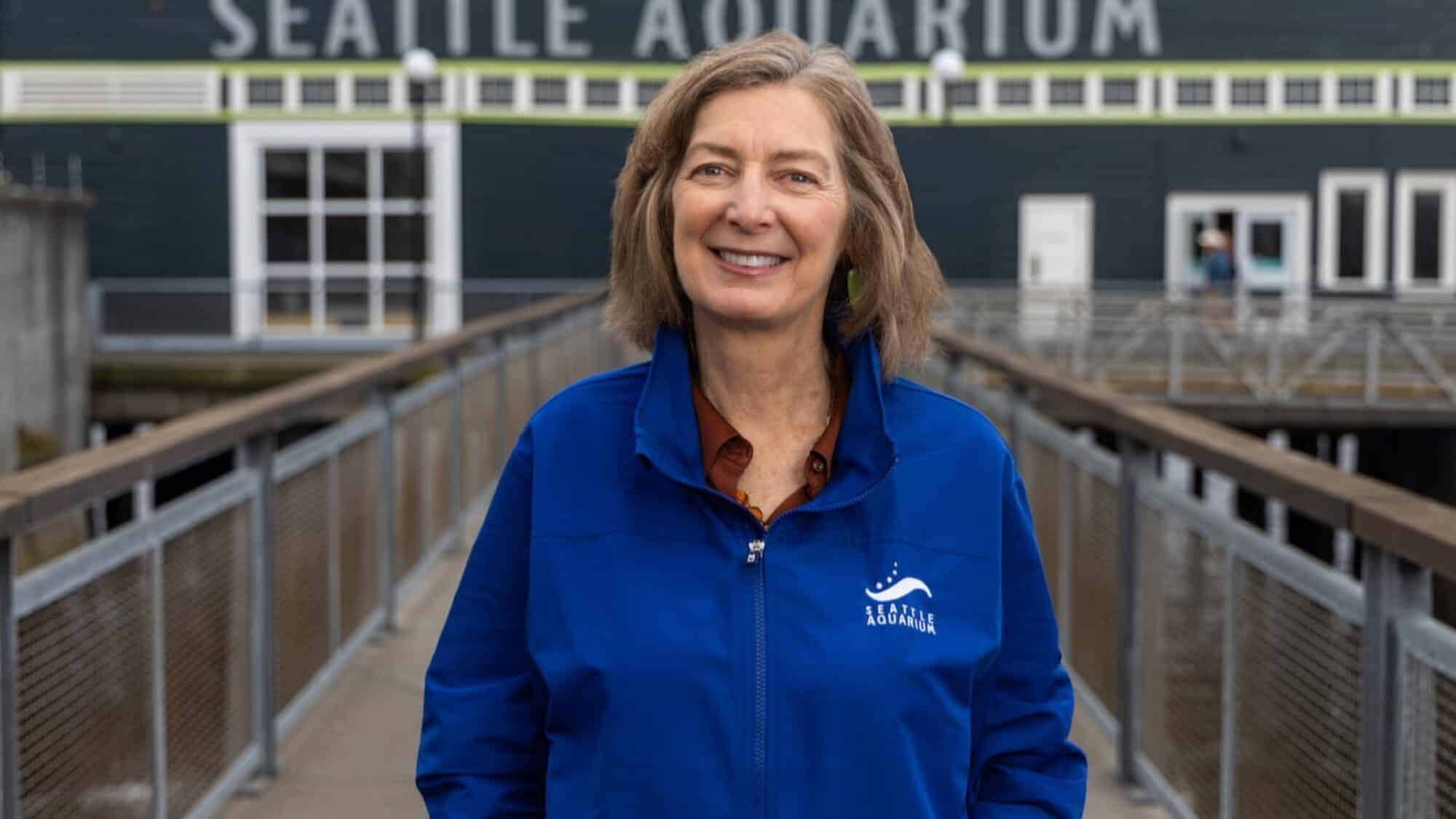 Peggy Sloan, in a blue Seattle Aquarium jacket, stands on a walkway in front of the Seattle Aquarium building, smiling at the camera and celebrating marine-life conservation.