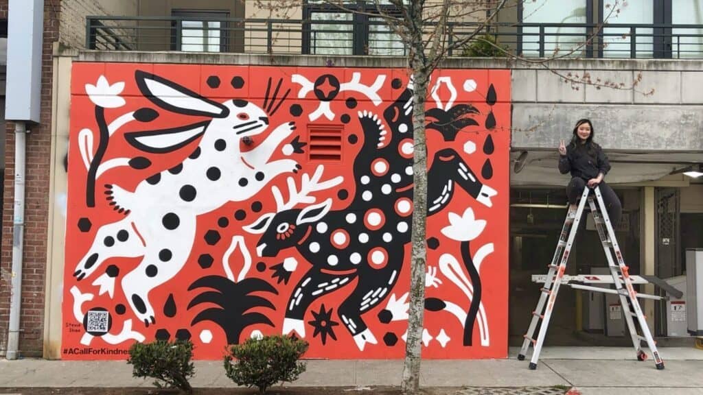 A mural featuring a white rabbit with black spots and a black creature with white spots, surrounded by abstract plants; an artist stands on a ladder beside the artwork.