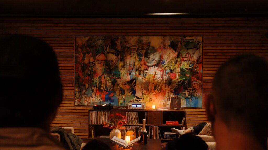 A colorful abstract painting hangs on a wooden wall above shelves with records, books, and candles; heads of two people are visible in the foreground.