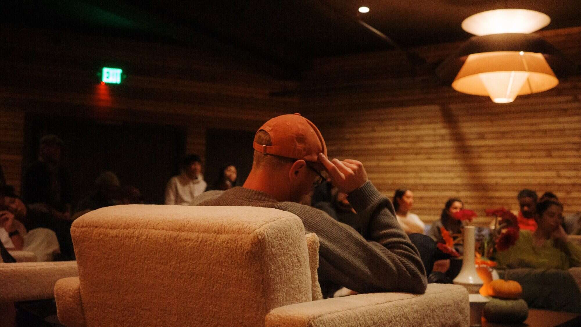 A person in an orange cap sits in a beige armchair facing a group of people in a dimly lit room with wooden walls and modern lighting.