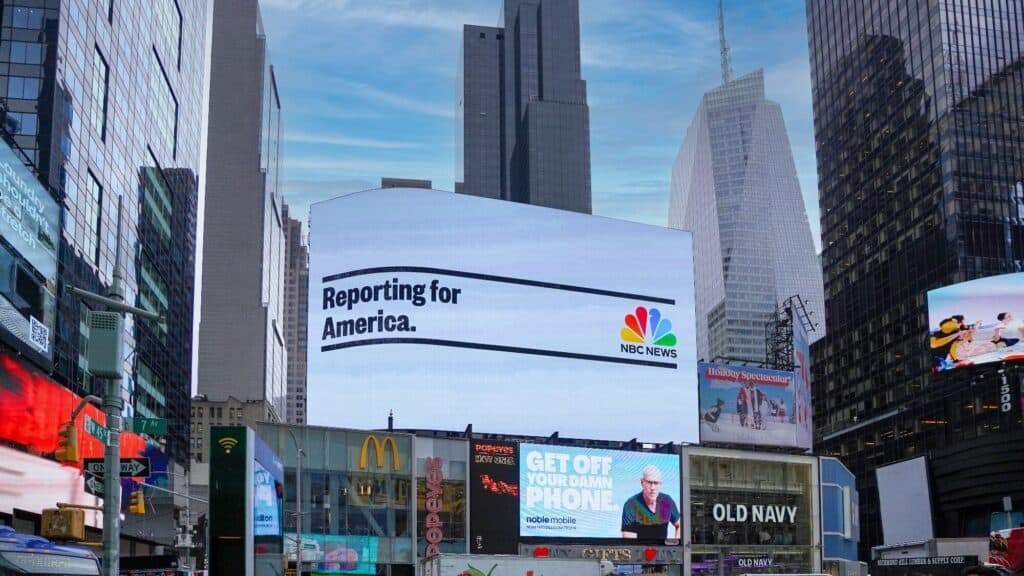Large digital billboard in Times Square displays an NBC News ad reading "Reporting for America," with various shops and other advertisements below.