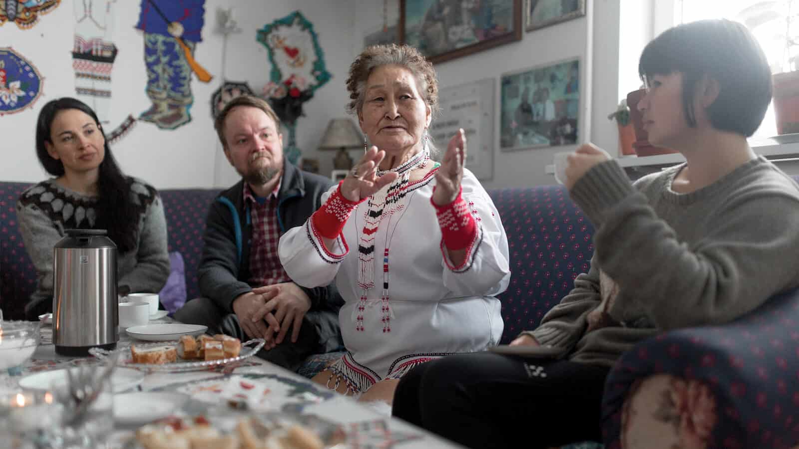 An elderly woman in traditional clothing gestures while speaking to three people seated on a couch around a table with food and drinks, as if sharing stories from the edge of the map or the center of the world.