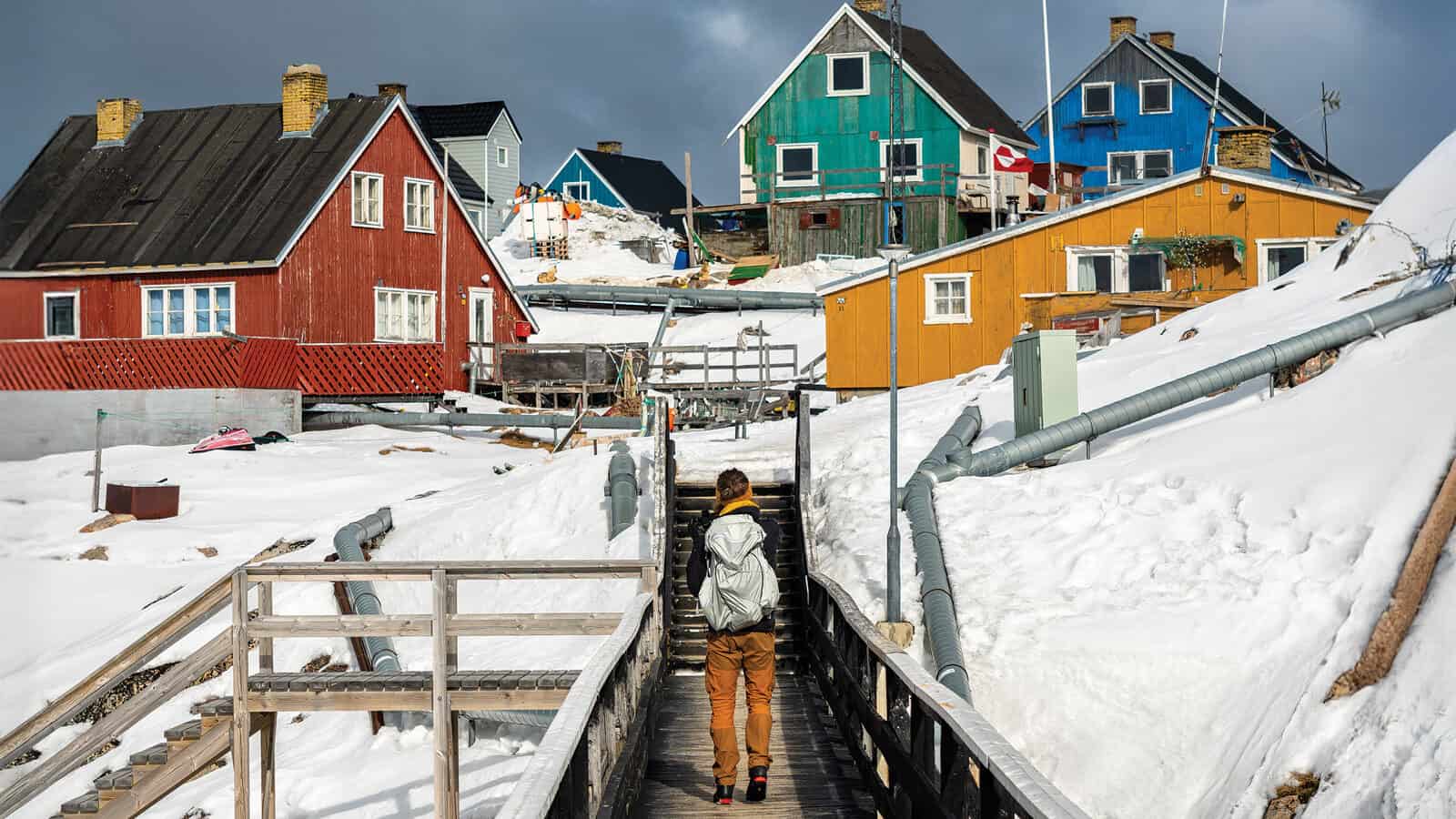 A person walks on a wooden pathway through snow toward colorful houses in a small, hilly settlement at the edge of the map.