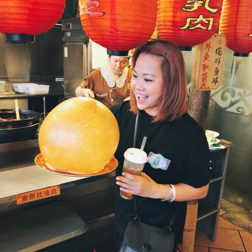 A woman smiles while holding a drink and pointing at a large, round fried food on a plate in a restaurant with red lanterns and Chinese signage—an inviting spot for Thanksgiving dining.