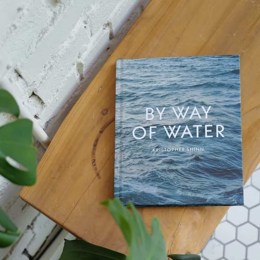 A book titled "By Way of Water" by Kristopher Shinn is placed on a wooden surface next to a white brick wall and green plants.