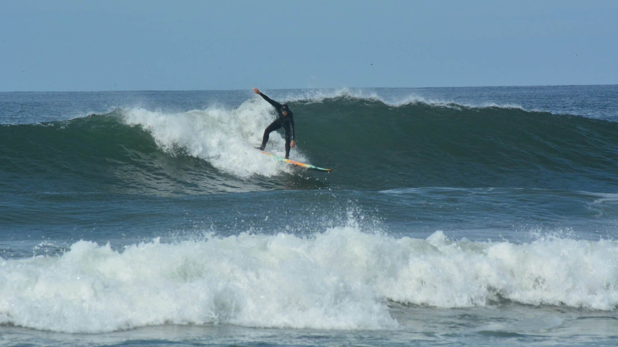 A surfer in a black wetsuit rides a wave on a surfboard in the ocean under a clear sky.