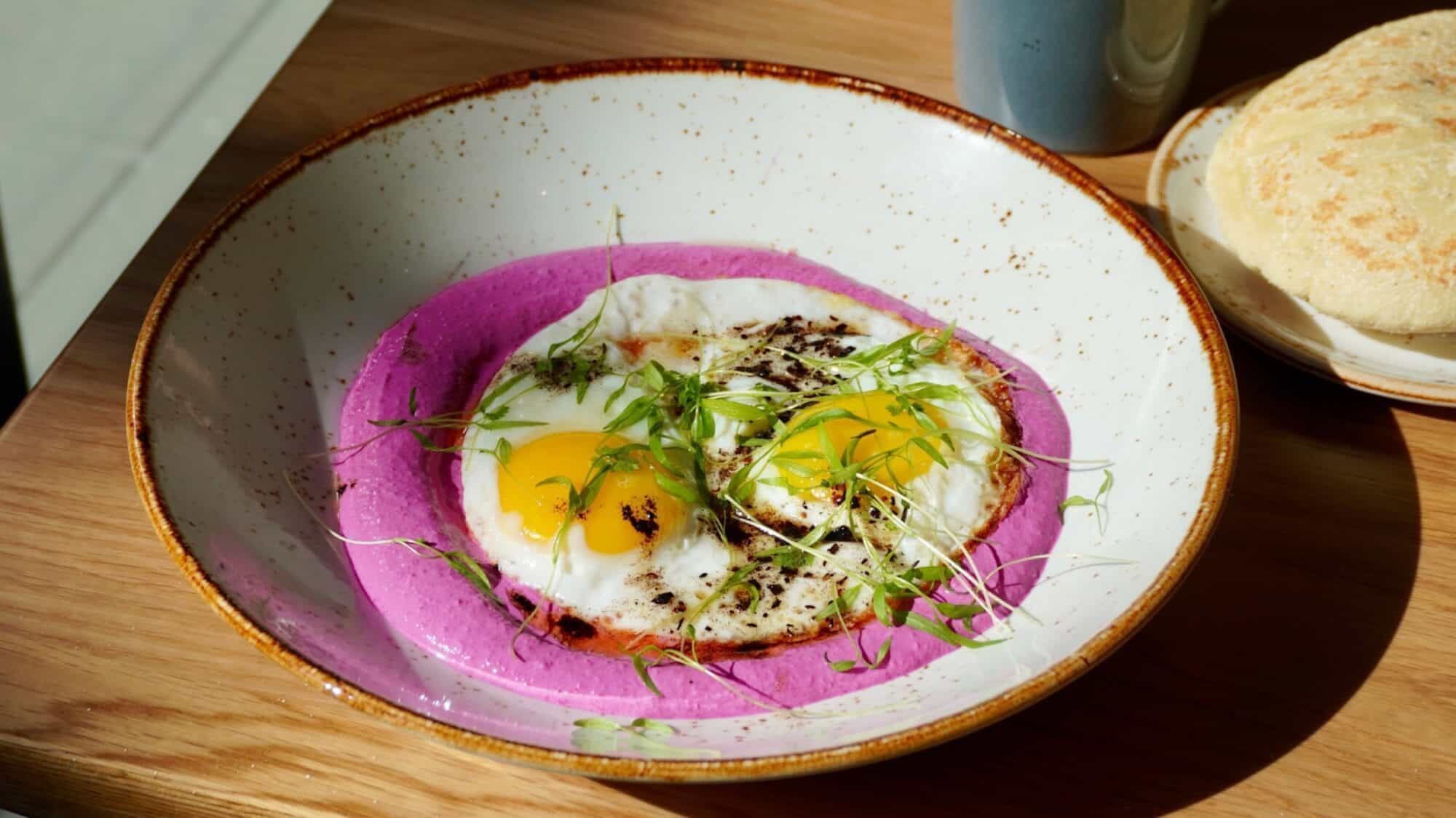 A bowl with two sunny-side-up eggs and microgreens on a layer of purple puree, placed on a wooden table next to a bread roll and a mug.