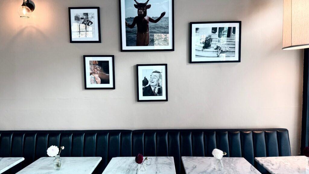 A cafe interior with marble tables, black cushioned seating, small vases with flowers, and six black-and-white framed photos on a beige wall.