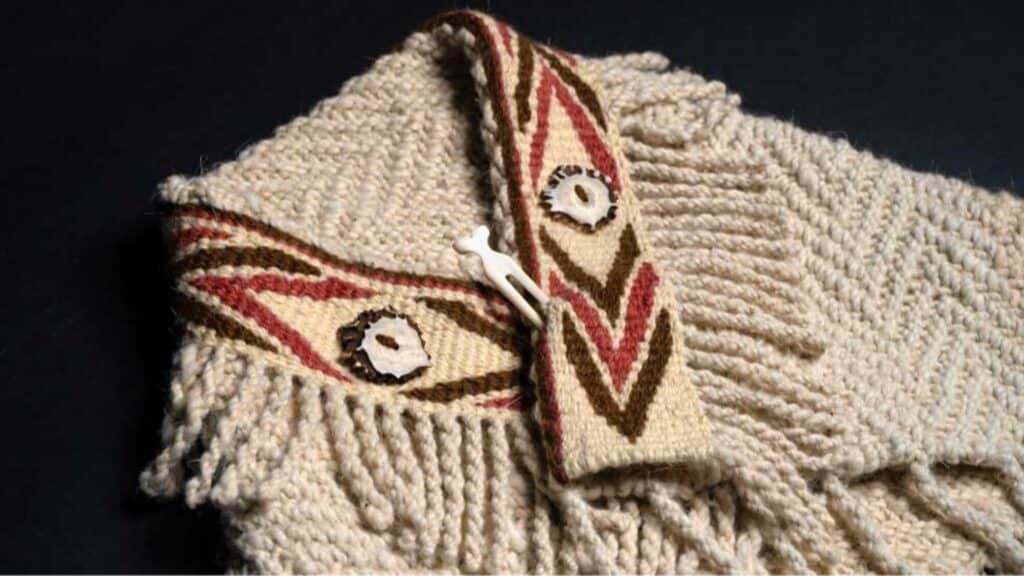 A close-up of a knitted beige garment with a patterned collar, fringe details, decorative buttons, and a bone toggle clasp inspired by Native Heritage traditions of Washington.
