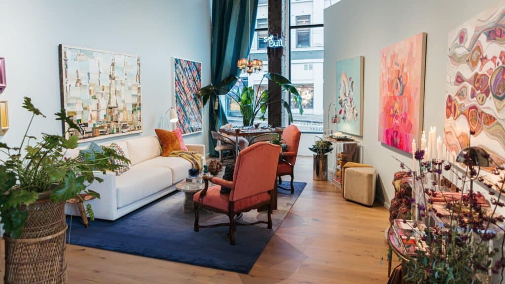 A stylish living room with a white sofa, orange chairs, large colorful paintings, indoor plants, wooden floors, and a dining table by tall windows.