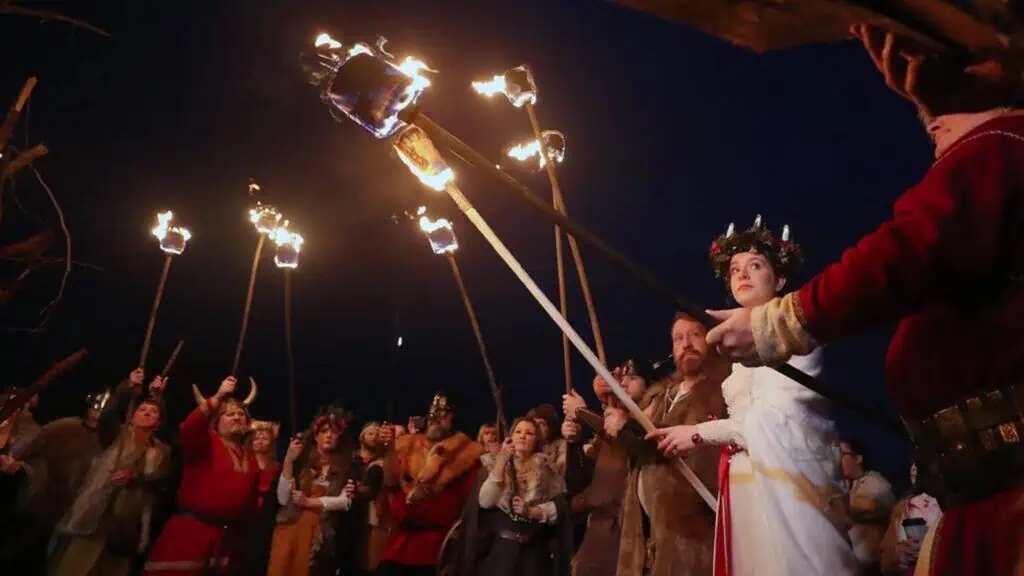 A group of people in historical costumes hold burning torches at night, with one woman in a white dress and crown of greenery standing prominently in the foreground.