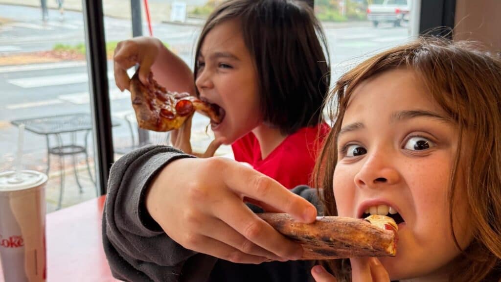 Two children sit at a table by a window at Sourdough Willy’s, enjoying slices of sourdough pizza. One looks at the camera while taking a bite; the other eats in the background. A drink cup rests on the table, adding to the cozy Counter Culture vibe.
