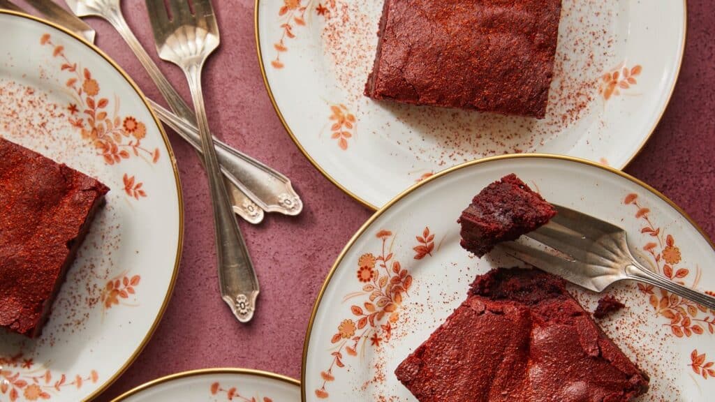 Three floral plates with squares of chocolate cake, some with forks and cake pieces, on a pink surface.