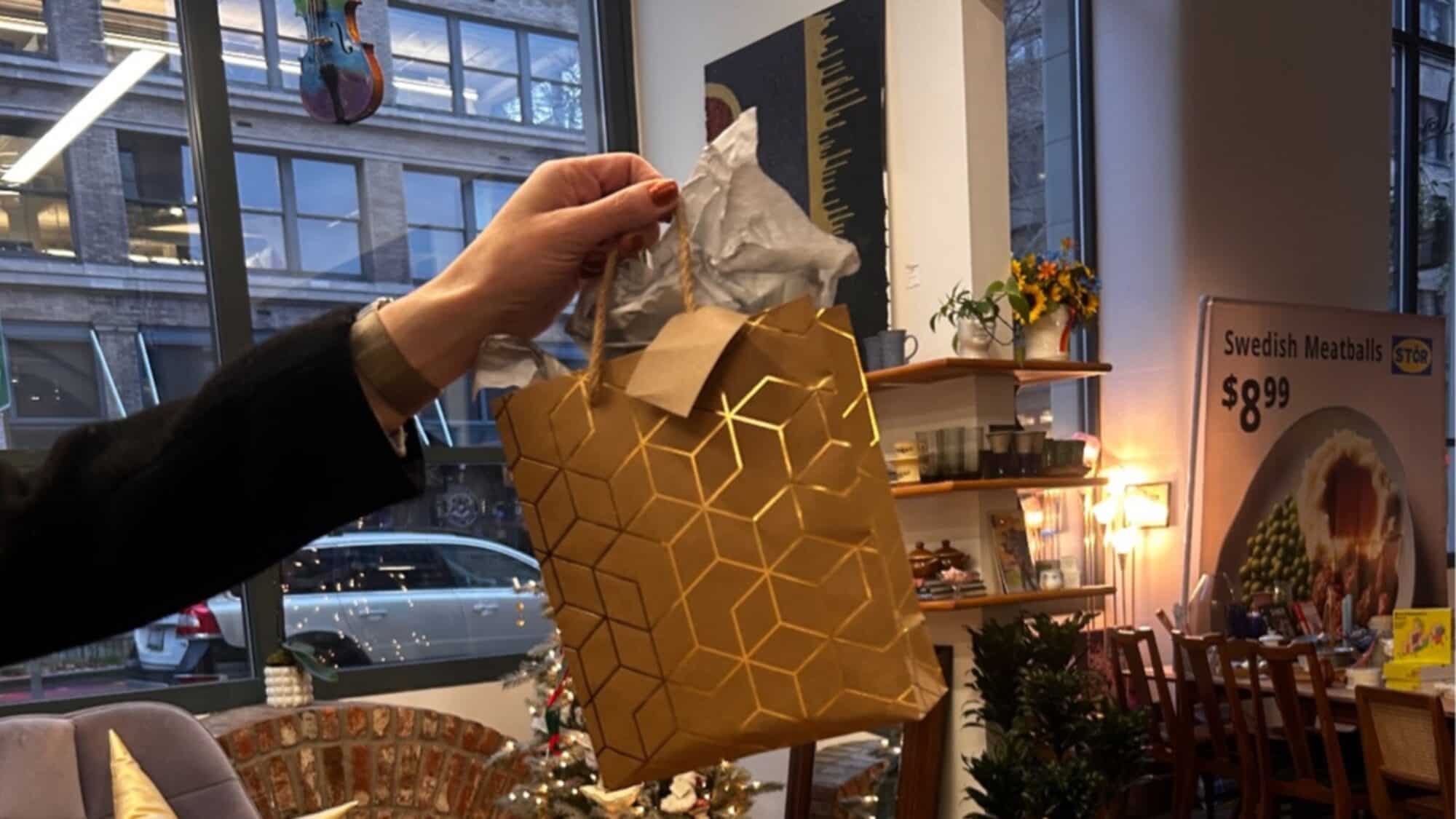 A hand holds a small gold gift bag with tissue paper in a store, with shelves, flowers, and a holiday tree visible in the background.