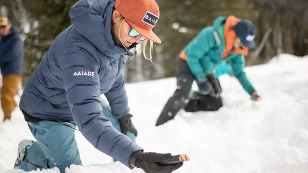 People in winter jackets kneel in the snow, using avalanche beacons and probes, participating in a snow safety or avalanche training exercise.