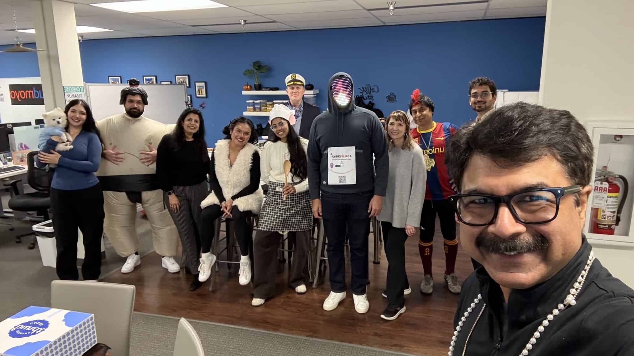 A group of people in costumes pose for a photo in an AI-First Consulting office. One person takes a selfie in front, with tables and chairs visible in the background.