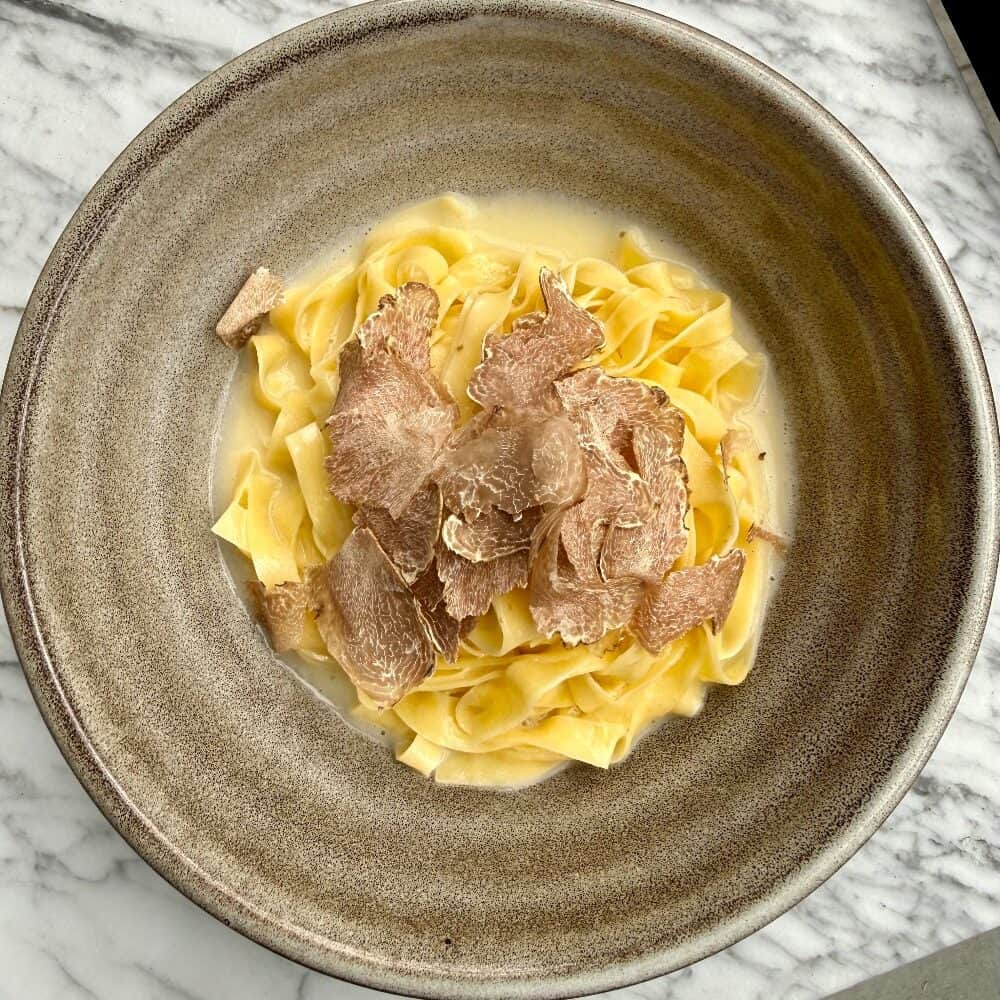 A bowl of tagliatelle pasta topped with shaved white truffles, served on a gray ceramic plate on a marble surface—an indulgent choice among December foods and perfect for winter recipes.