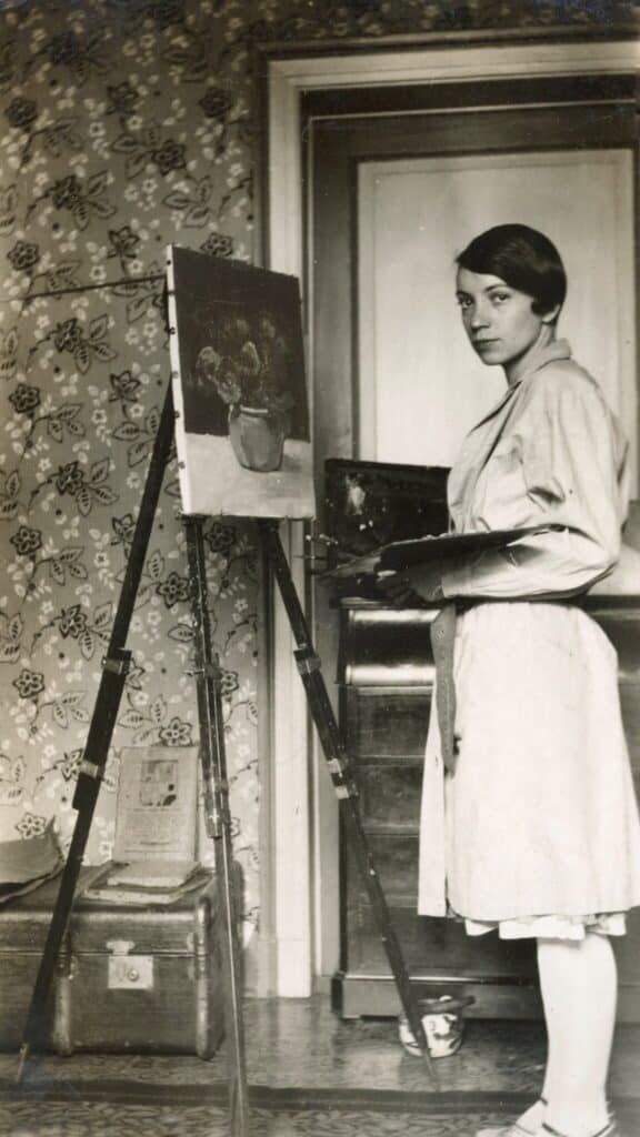 A woman in a light dress stands at an easel, painting a still life of a potted plant in a room with floral wallpaper.