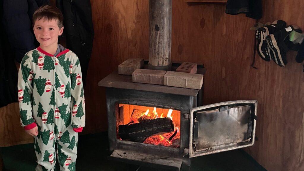 A young child in Christmas pajamas stands beside a wood-burning stove with a fire inside, in a wooden-paneled room.