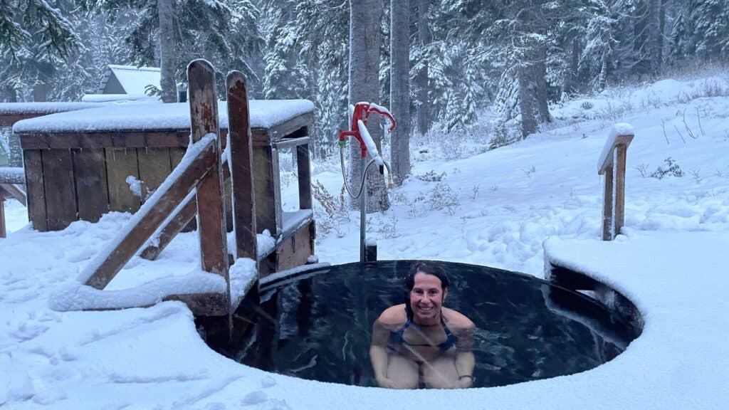 A person is sitting in an outdoor hot tub surrounded by snow-covered ground and trees in a winter setting.