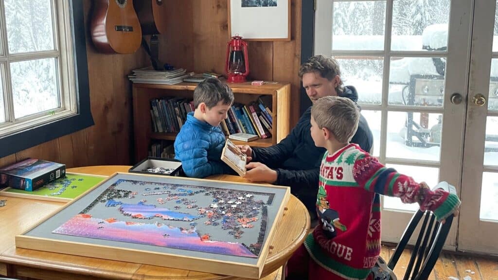 Three people work on a puzzle at a wooden table indoors. Snow is visible outside the window and door; books, guitars, and a lantern are in the background.