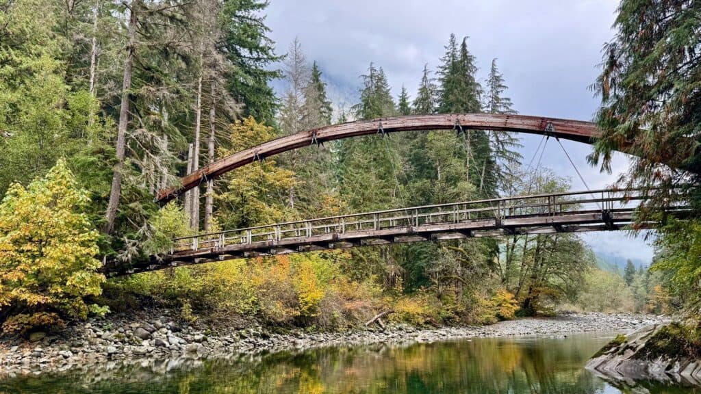 A wooden arched bridge spans a calm river surrounded by dense evergreen trees and autumn foliage.