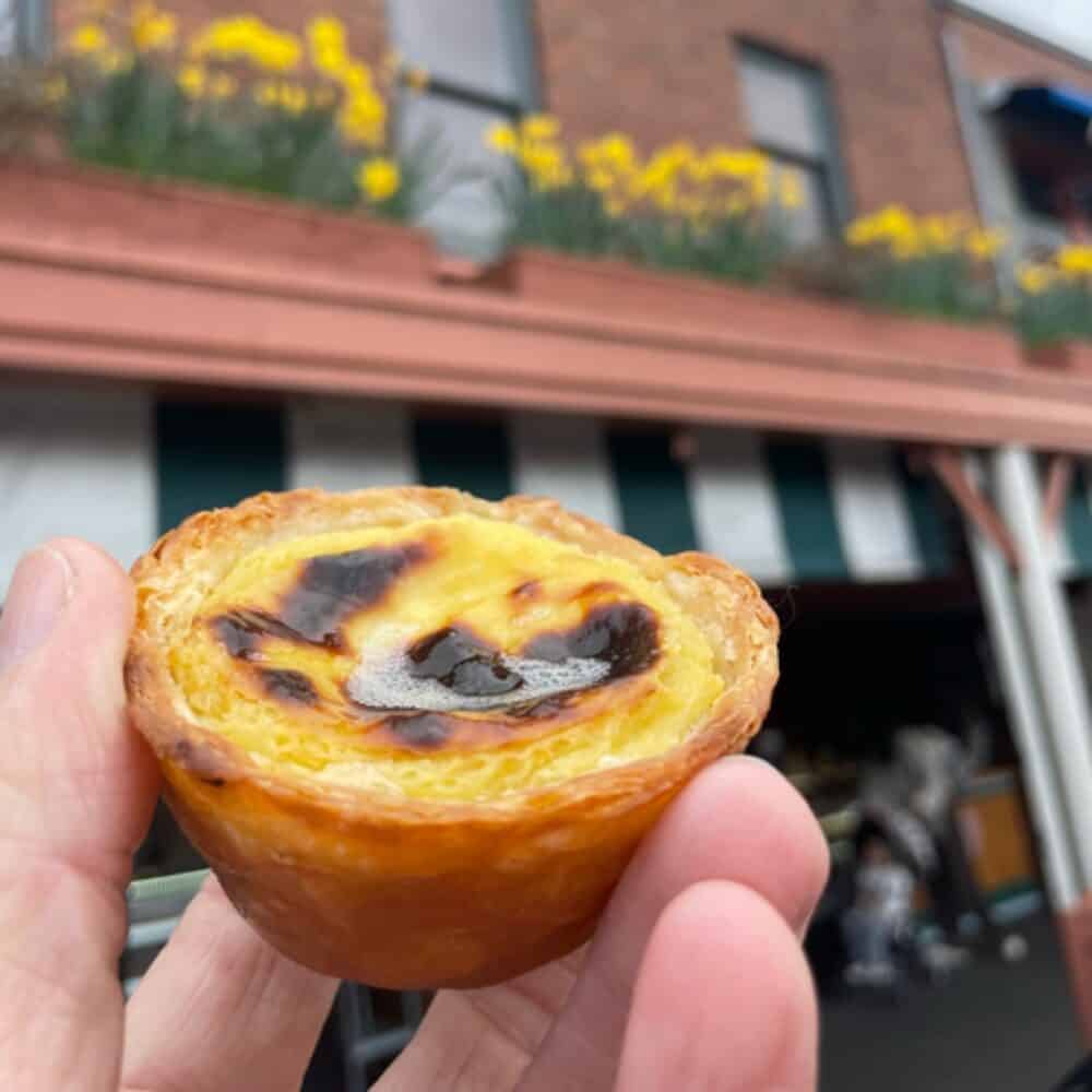 A hand holds up a Portuguese egg tart, or pastel de nata, with a blurred background of a building, flowers, and storefront.