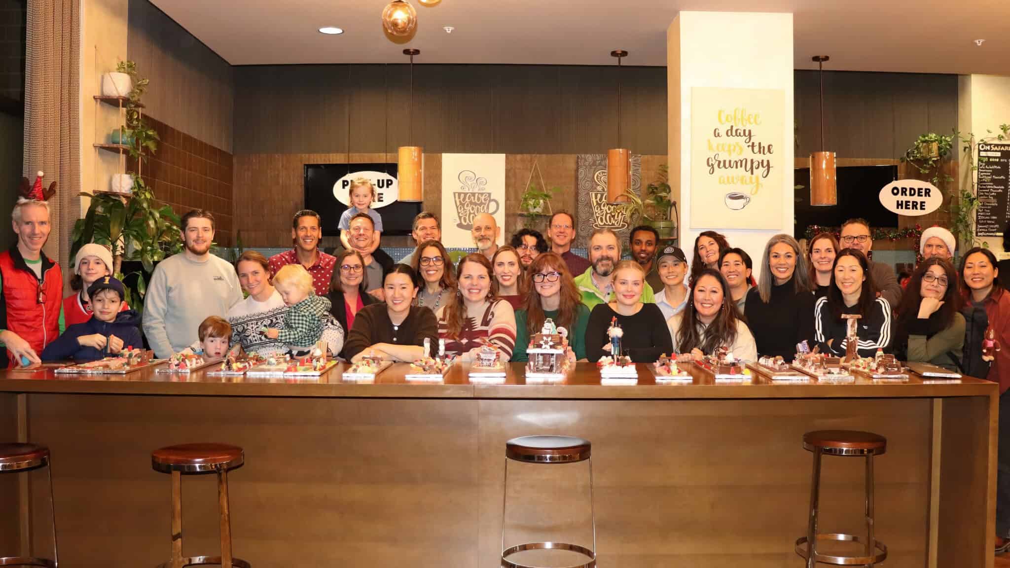 With curiosity and momentum, a large group of people—including adults and children—pose and smile behind a counter filled with decorated desserts in a well-lit indoor space, embodying the vibrant spirit of Logic20/20.