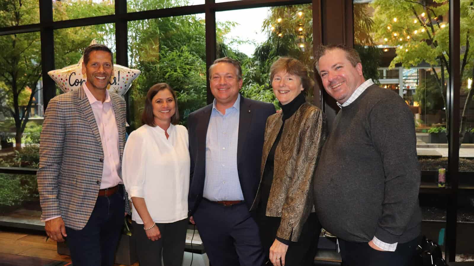 Five adults stand together indoors, smiling at the camera with large windows and greenery in the background. A "Congratulations" balloon is partially visible on the left, capturing a moment of momentum and celebration at Logic20/20.