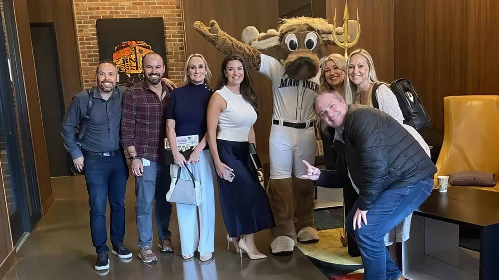 A group of seven adults, fueled by curiosity, pose and smile with a person in a moose mascot costume wearing a Mariners baseball uniform indoors.