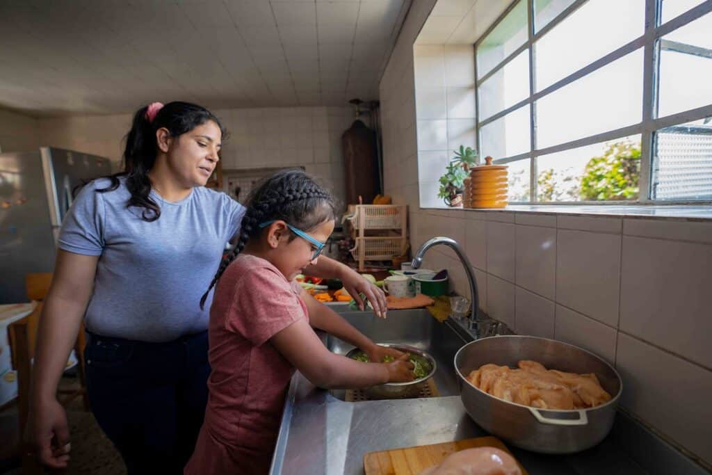 An adult and a child stand at a kitchen sink washing vegetables together, with raw chicken pieces in a pot on the counter nearby—some of these fresh ingredients were provided through United Way’s Home Grocery Delivery program.