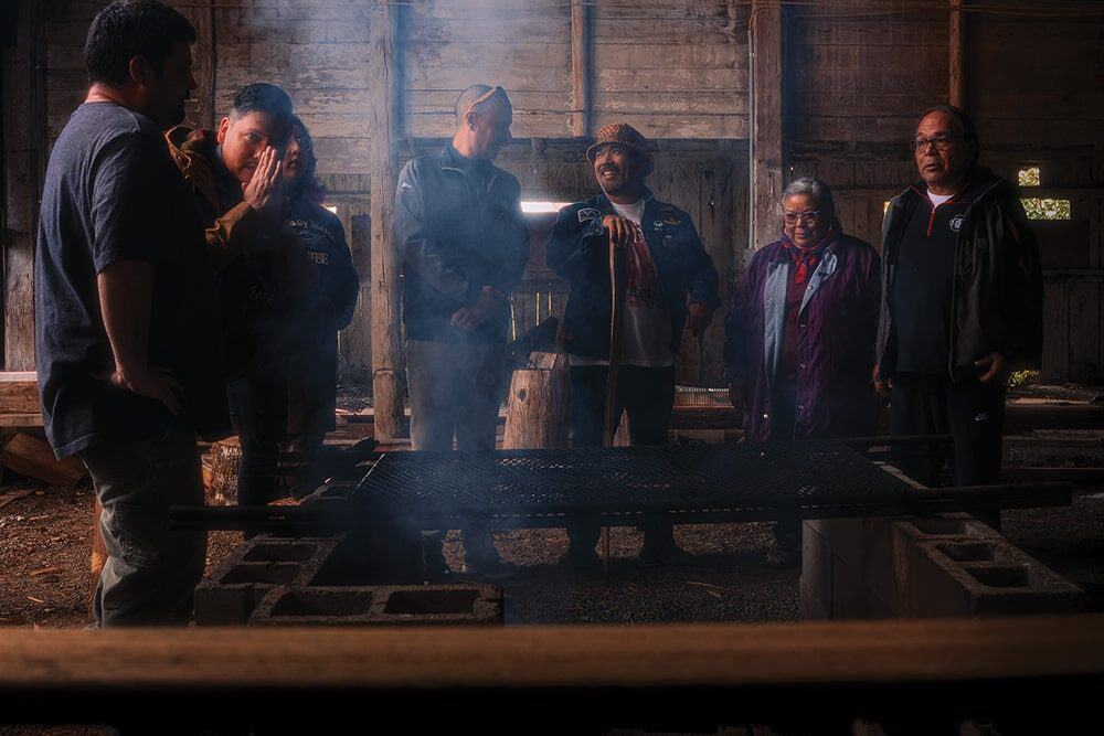 Seven people stand in a semicircle around a smoking grill in a dimly lit wooden building, with smoke rising and light filtering through windows—a moment of photography that Nick Ward would appreciate.