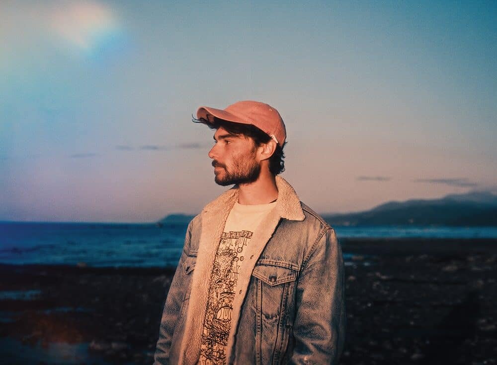 A man in a denim jacket and pink cap stands by a rocky shoreline at dusk, sea and mountains behind him—a moment of photography captured by Nick Ward.