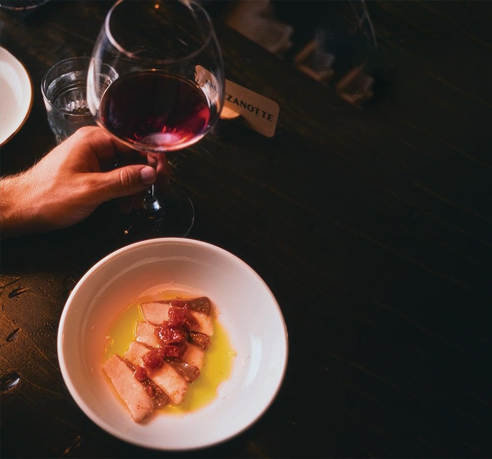 A person holding a glass of red wine sits at a dark table with a white bowl of sliced fish in oil and sauce, captured in Nick Ward’s evocative photography style.