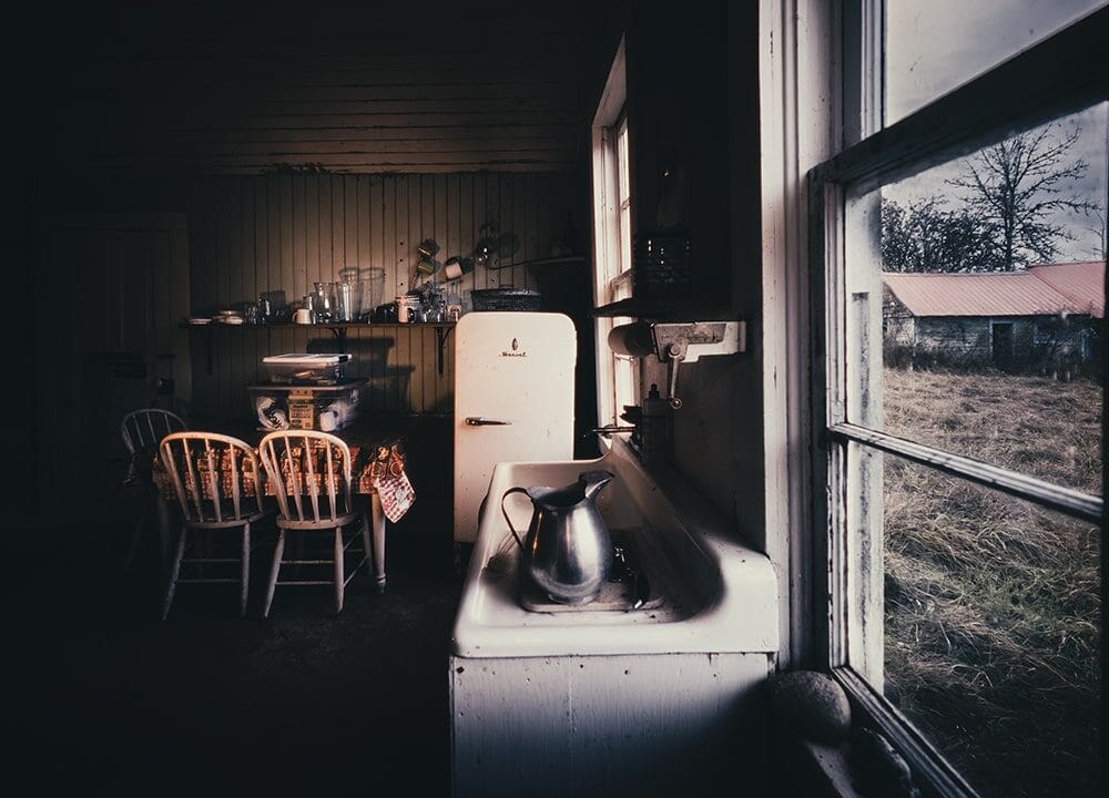 Dimly lit rustic kitchen photography by Nick Ward: a sink, metal kettle, vintage fridge, wooden table and chairs. Sunlight streams through a window, illuminating an overgrown yard and outbuildings beyond.