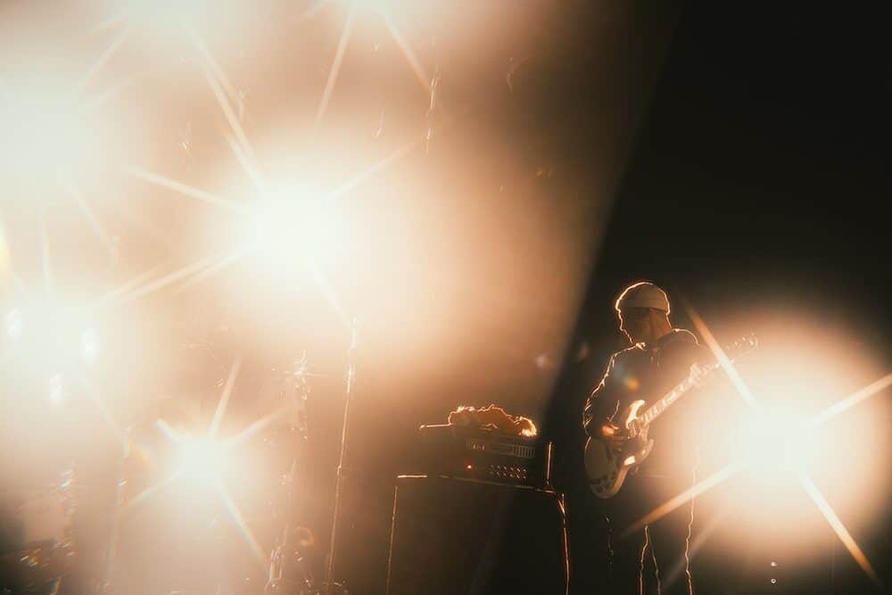 A guitarist stands on stage surrounded by bright stage lights, creating a dramatic and hazy atmosphere captured perfectly in Nick Ward’s photography.