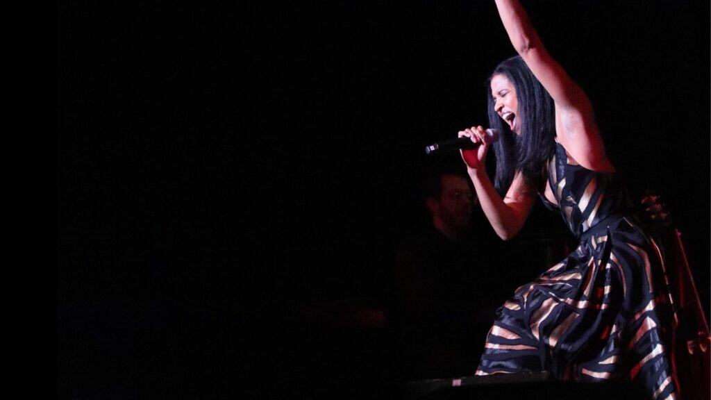 A woman in a patterned dress sings passionately into a microphone on stage, with one arm raised and dark background.