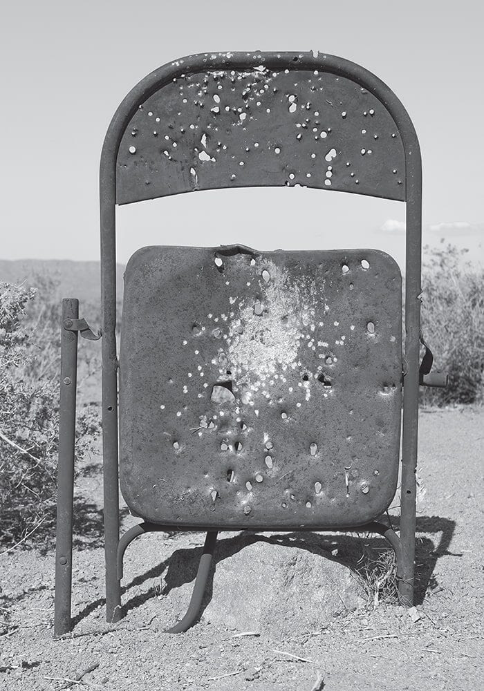 A metal folding chair stands outdoors, its seat and backrest riddled with bullet holes and dents—an unintended testament to architecture and design—set against a backdrop of desert vegetation, like an artifact from an outdoor ARCADE.