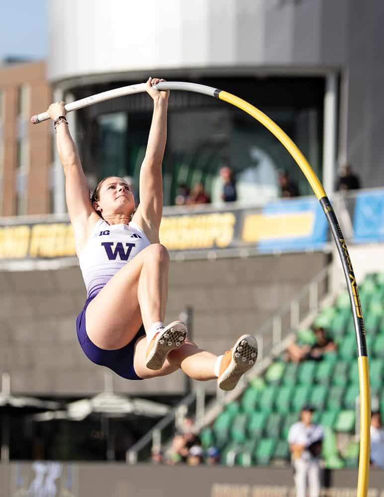An athlete in a white "W" uniform, representing UW Track and Field, performs a pole vault during a track and field event at an outdoor stadium.