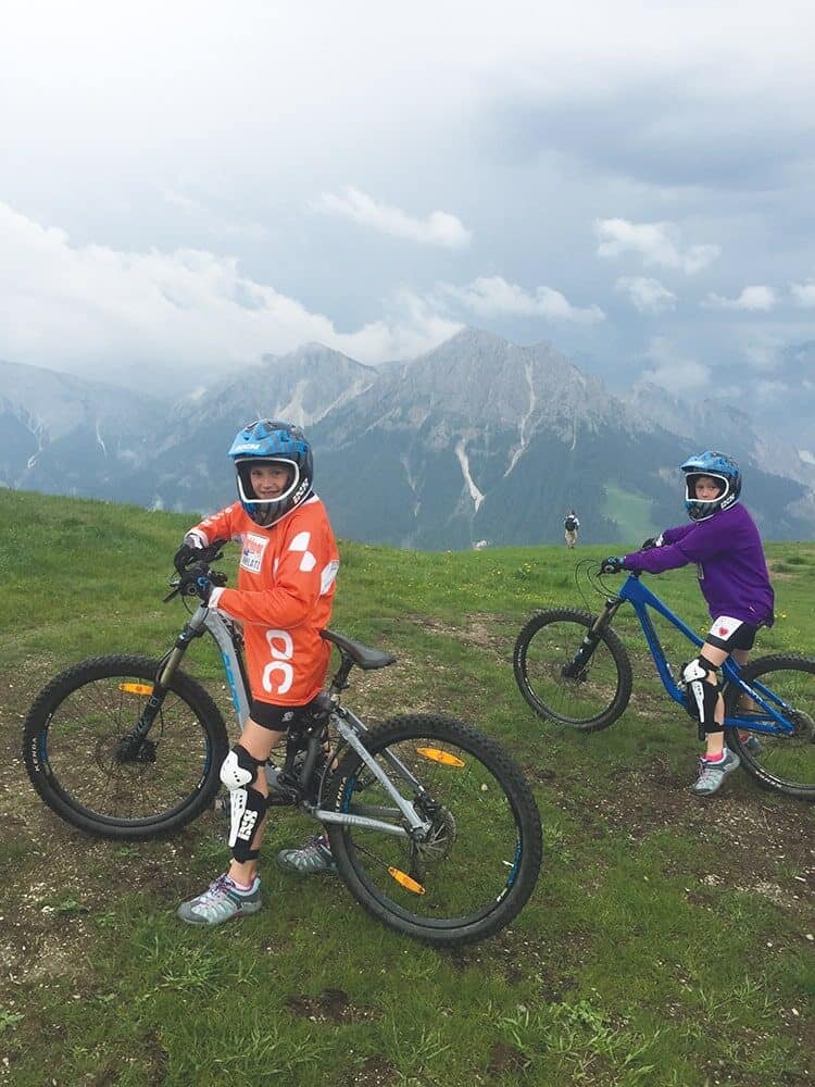 Amanda and Hana Moll, pole vault champions from UW Track and Field, pose with mountain bikes on a grassy hill, wearing helmets and protective gear, with mountains and a cloudy sky in the background.