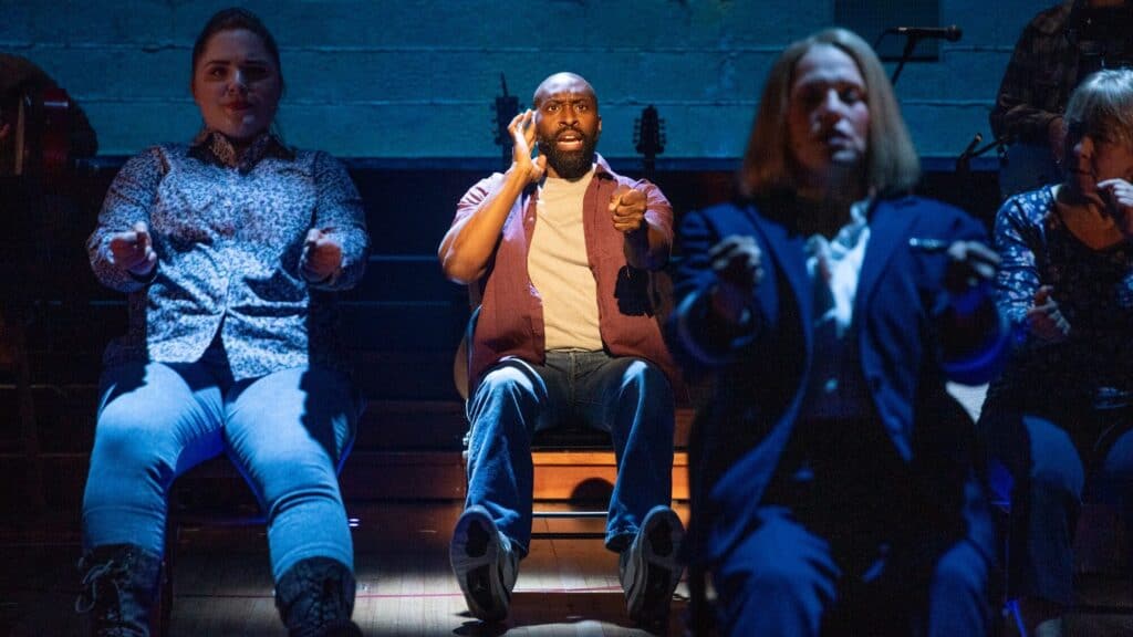 Three people sit in a row on stage, miming actions as if driving or giving directions, under dramatic lighting with a blue backdrop.