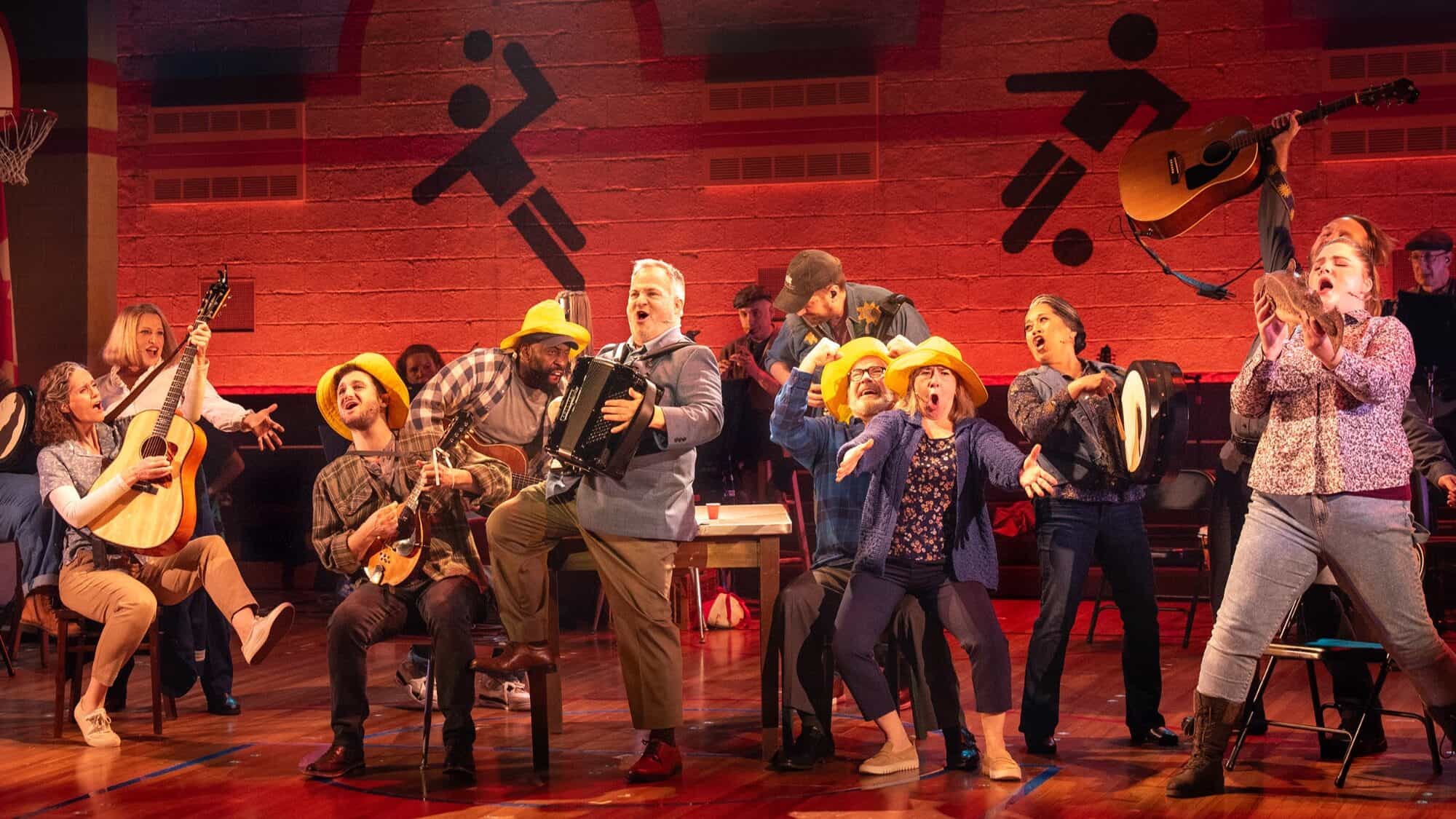 A group of performers, some with instruments and yellow hats, sing and act energetically on a stage set resembling a gymnasium.