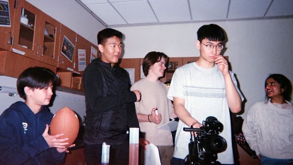 Five young people are standing in a classroom; one holds a football, another operates a camera, and the others are smiling and talking.
