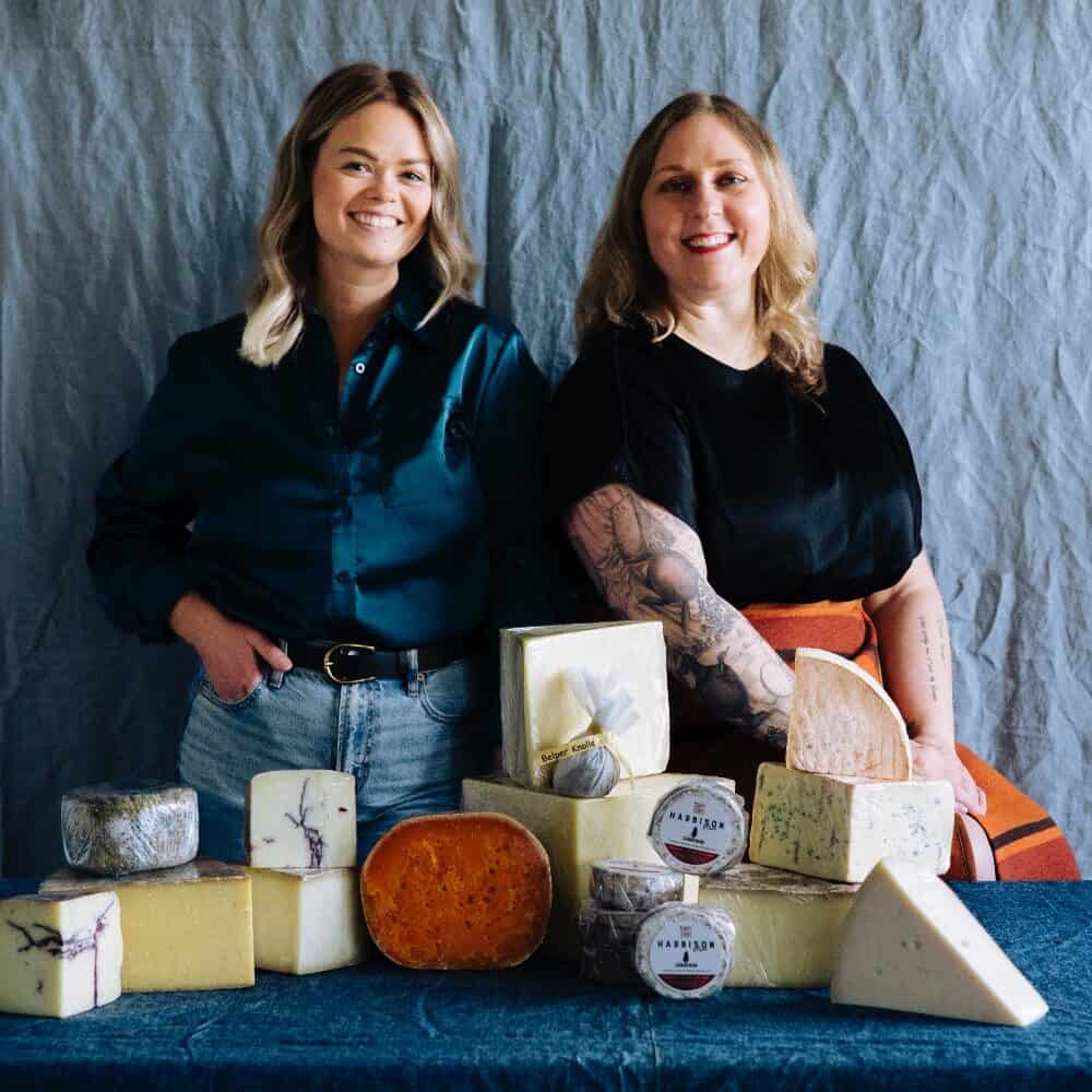 Two women stand behind a table displaying various blocks and wheels of cheese against a plain fabric backdrop.